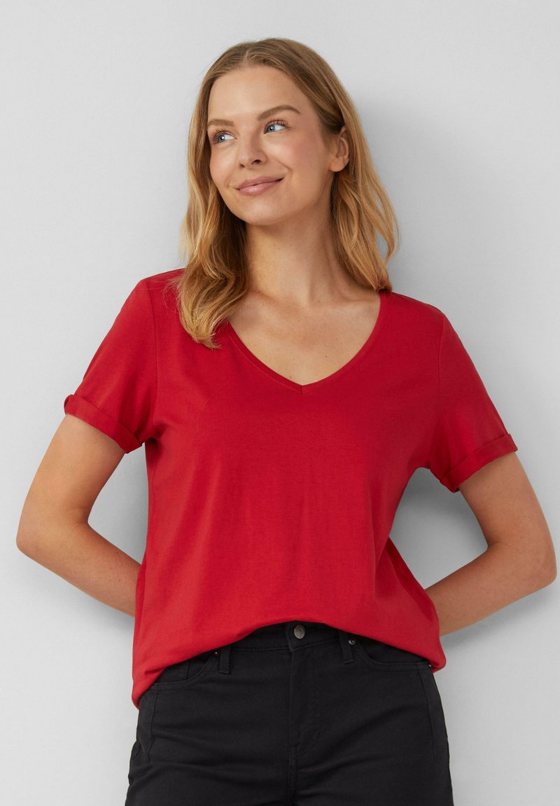 T-shirt rossa a maniche corte con scollo a V, realizzata in un tessuto morbido. Maniche arrotolate e vestibilità comoda. Abbinata a pantaloni neri.