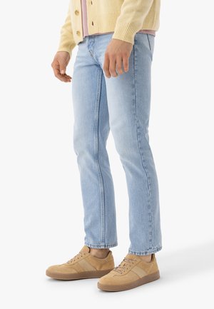 Osoba ubrana w jasnoniebieskie dżinsy, beżowe trampki oraz bladoróżowy rozpinany kardigan, stojąca na białym tle.