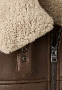 Giacca di pelle marrone con un colletto in shearling soffice di colore crema, accessori in metallo argentato e una cerniera con linguetta brandizzata.