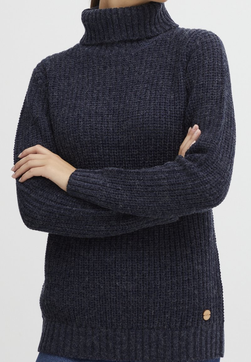 Personne portant un pull à col roulé en maille épaisse bleu foncé, les bras croisés, montrant la texture et les détails des bordures côtelées.