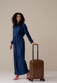 Navy pleated jumpsuit features long sleeves and cinched waist. Brown hard-shell suitcase with ridged texture and four wheels; black telescopic handle.
