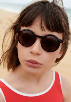 Ochelari de soare rotunzi, întunecați, cu rame groase și o ușoară formă cat-eye; purtați de o persoană cu păr șaten deschis, îmbrăcată într-un costum de baie roșu cu margine albă.