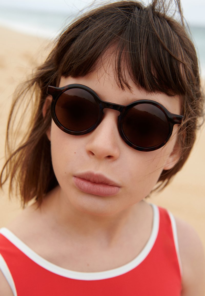 Occhiali da sole rotondi scuri con montatura spessa e leggermente a forma di occhio di gatto; indossati da una persona con capelli castano chiaro, che indossa un costume da bagno rosso con bordi bianchi.