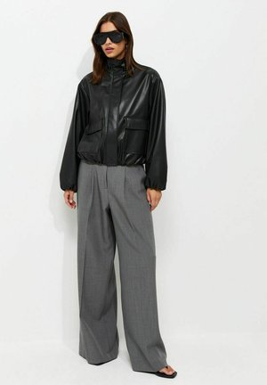 Donna che indossa una giacca di pelle nera oversize, pantaloni grigi a gamba larga, occhiali da sole neri e sandali neri, in piedi su uno sfondo bianco.