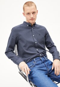 Navy-blau gestreiftes Hemd mit Button-Down-Kragen und langen Ärmeln, kombiniert mit blauen Jeans. Subtiler Logo-Detail auf dem Hemd. Glatte Stofftextur.
