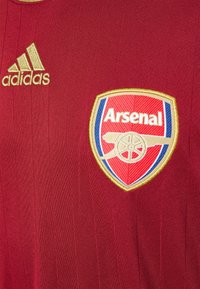 Czerwona koszulka z pionowymi paskami, wykonana z lekkiego materiału. Posiada złote logo Adidas oraz haftowany herb Arsenalu z czerwonymi, niebieskimi i złotymi detalami.