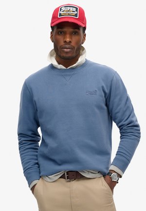 Lichtblauwe crewneck sweatshirt met geribbelde mouwen, over een grijs shirt. Een rode pet met een witte patch waarop "Super Racing Team" staat. Bruine riem zichtbaar.