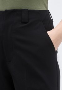 Άτομο με μαύρο παντελόνι, το οποίο έχει το χέρι του στην δεξιά τσέπη και εμφανή θηλιά ζώνης, συνδυασμένο με πράσινο ελιάς τοπ.