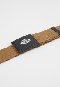Tan vävt bälte gjort av tyg med en texturerad yta. Har ett svart metallspänne med "Dickies"-logotypen tydligt synlig.