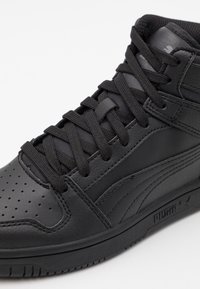 Černá vysoká teniska s perforovanou špičkou, hladkou kůží a textilními materiály, plochými černými tkaničkami a minimalistickými designovými akcenty.