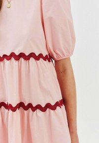 Różowa bawełniana sukienka z bufiastymi rękawami, zdobiona falbankami w kolorze czerwonym wzdłuż gorsetu i u dołu, tworząca teksturowany, warstwowy wygląd.