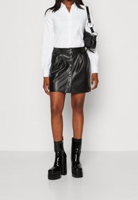 Jupe mini en cuir noir avec fermeture à boutons à l'avant, taille haute et plis subtils ; associée à une chemise blanche à boutons parfaitement inspirée et des bottes à plateforme noires.