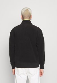 Svart huvjacka med hög krage, med en gul accent inuti kragen, tillverkad av mjukt, strukturerat tyg. Avslappnad passform.