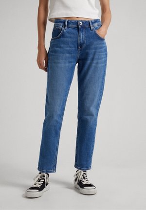 Personne portant un jean taille mi-haute bleu à jambes droites et des baskets noir et blanc, debout avec une main dans la poche.