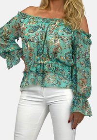 Turkos off-shoulder blus med blommig paisleymönster, volangärmar och rynkad midja. Kombinerat med vita byxor.