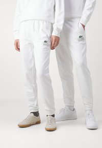 Bílé teplákové kalhoty s elastickým pasem a manžetami, se zeleným logem Lacoste. Vytvořeno v kombinaci se světle béžovými a bílými botami.