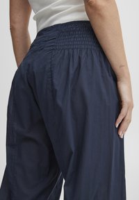 Naine kannab tumesiniseid lahtisi pükse, millel on kogutud elastne vöökoht, ja valget varrukateta topi, seisab ühevärvilise tausta ees.