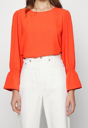 Femme portant un chemisier orange vif à manches longues avec des poignets évasés et un jean blanc taille haute, debout devant un fond clair.
