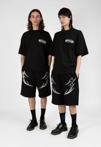 Black t-shirts with logo, paired with black shorts featuring silver flame patterns. Both wear black socks and shoes. Unisex design.