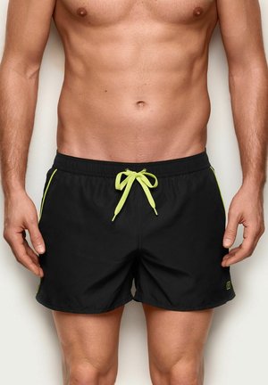 Mężczyzna w czarnych kąpielówkach z neonowo-żółtym sznurkiem i lamówką, stojący na jednolitym jasnym tle.