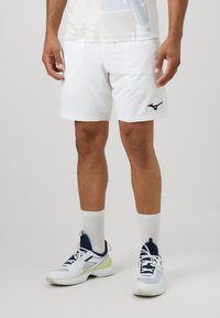 Weiße Sportshorts mit einem schwarzen Logo auf dem linken Bein, kombiniert mit weißen Socken und weißen-blauen Sportschuhen mit grünen Akzenten.
