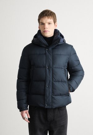 Doudoune marine avec fermeture éclair et à boutons, capuche, texture matelassée et poches latérales zippées. Portée par-dessus un col roulé noir.