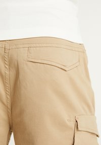 Shorts en khaki avec une poche arrière plate et une poche cargo sur le côté, fabriqués à partir d'un tissu léger, présentant des coutures surpiquées et une texture lisse.