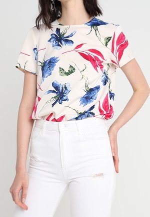 Woman wearing a white short-sleeve top with blue and red floral prints paired with high-waisted white jeans with ripped details.
