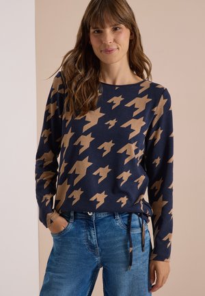 Femme aux longs cheveux bruns porte un pull bleu marine avec un grand motif oiseau beige et un jean bleu, debout avec une main dans la poche.