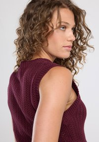Bordeaux neulepusero ilman hihoja, jossa on V-pääntie, teksturoitu kuvio ja ribbireunat. Keskipitkä malli, tiukka siluetti.