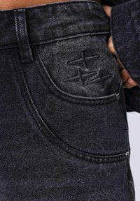 Jeans de mezclilla negra con un primer plano de un bolsillo delantero con adornos bordados y un detalle de cremallera distintivo.