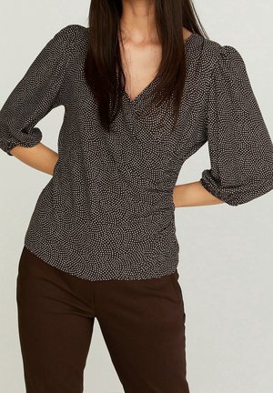 Kobieta w brązowej bluzce w groszki z dekoltem w kształcie litery V, z rękawami o długości do łokcia z bufkami, oraz w ciemnobrązowych spodniach, z rękami spoczywającymi na biodrach.