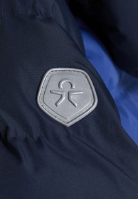 Jūros mėlynos spalvos audinys su lygia tekstūra, turintis sidabrinį emblemą ant skydelio formos paškės, demonstruojantis paprastus dizaino elementus.