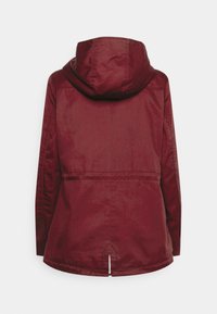 Veste imperméable bordeaux avec capuche, tissu doux, coupe ajustée et une fente au dos pour la respirabilité. Coutures uniques surpiquées.