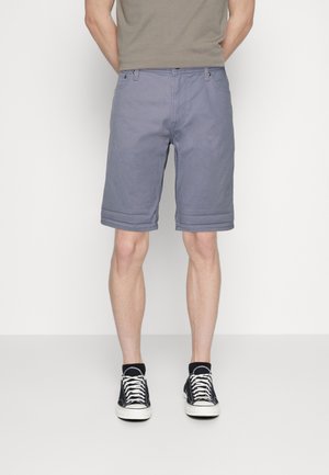 Mann trägt graue knielange Shorts, schwarz-weiße Sneakers, steht mit den Händen hinter dem Rücken vor einfarbigem Hintergrund.