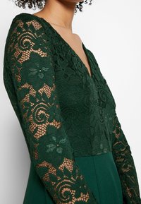 Grünes Kleid mit V-Ausschnitt, langen Ärmeln aus floraler Spitze und einem glatten, taillierten Oberteil sowie einem fließenden unteren Teil.