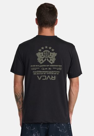 Mężczyzna z krótkimi ciemnymi włosami pokazuje tył czarnej koszulki z grafiką dwóch lwów, globusa, gwiazd oraz napisem zawierającym "RVCA" i współrzędne.