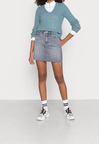 Grauer Denim-Minirock mit fransigem Saum, kombiniert mit einem türkisfarbenen Strickpullover und einem weißen Hemd mit Kragen, abgerundet mit schwarz-weißen Turnschuhen.