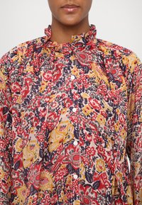 Blomstermønstret bluse med flæsekrave, prydet med røde, gule og marineblå farver på en lys baggrund, designet med knapper nedad foran.