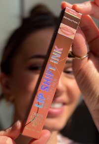 Lip Shift Ink matita per labbra in un tubo corallo-arancio con testo olografico. Presenta l'etichetta "Nude Talk", una forma rettangolare elegante e un design peel-off.