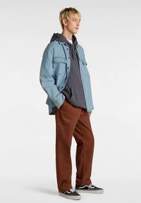 Jasnoniebieska kurtka dżinsowa na szarej bluzie z kapturem, brązowe spodnie i czarne trampki. Kurtka posiada dwie przednie kieszenie i luźny krój.