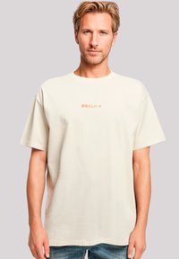 Beige katoenen t-shirt met korte mouwen, voorzien van een klein rood tekstontwerp in het midden. Eenvoudige, ontspannen pasvorm met een ronde hals.