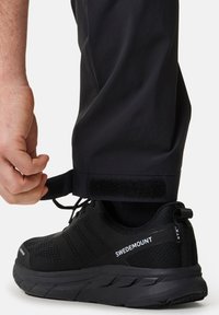 Chaussure de sport noire avec tige en mesh et semelle en caoutchouc. Une sangle en Velcro visible est en cours d'ajustement au niveau de la cheville. L'étiquette de la marque indique "SWEDEMOUNT."