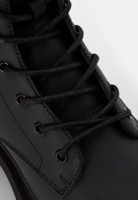 Bota de cuero negro con cordones negros redondos, ojales de metal, costura y una superficie texturizada. Enfoque en la sección superior de la bota.