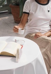 Camiseta blanca con ribetes negros y un pequeño gráfico de corazón, falda beige, sosteniendo una taza de café blanca al lado de un libro abierto sobre una mesa redonda blanca.