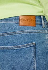 Niebieskie dżinsy Wrangler z brązową skórzaną naszywką w talii, z widocznym przeszyciem i szczegółami tylnej kieszeni.