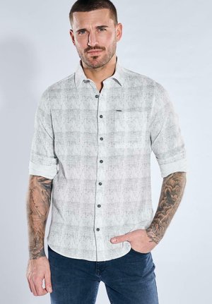 Mann mit kurzen Haaren und Tattoos trägt ein hellgraues Hemd mit aufgerollten Ärmeln und dunkelblaue Jeans, steht mit einer Hand in der Hosentasche.