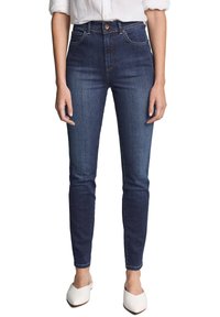 Jeans skinny azul oscuro de denim, con una cintura alta, costuras sutiles y diseño de cinco bolsillos. Combinados con zapatillas blancas tipo slip-on.