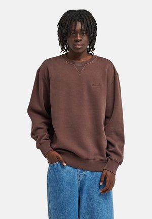 Giovane con dreadlocks alle spalle che indossa una felpa marrone e jeans blu, in piedi con una mano in tasca su uno sfondo semplice.