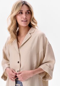 Beżowa koszula z guzikami, o luźnym kroju, z oversize'owymi rękawami i miękką fakturą, z klasycznym kołnierzykiem i ciemnymi guzikami.